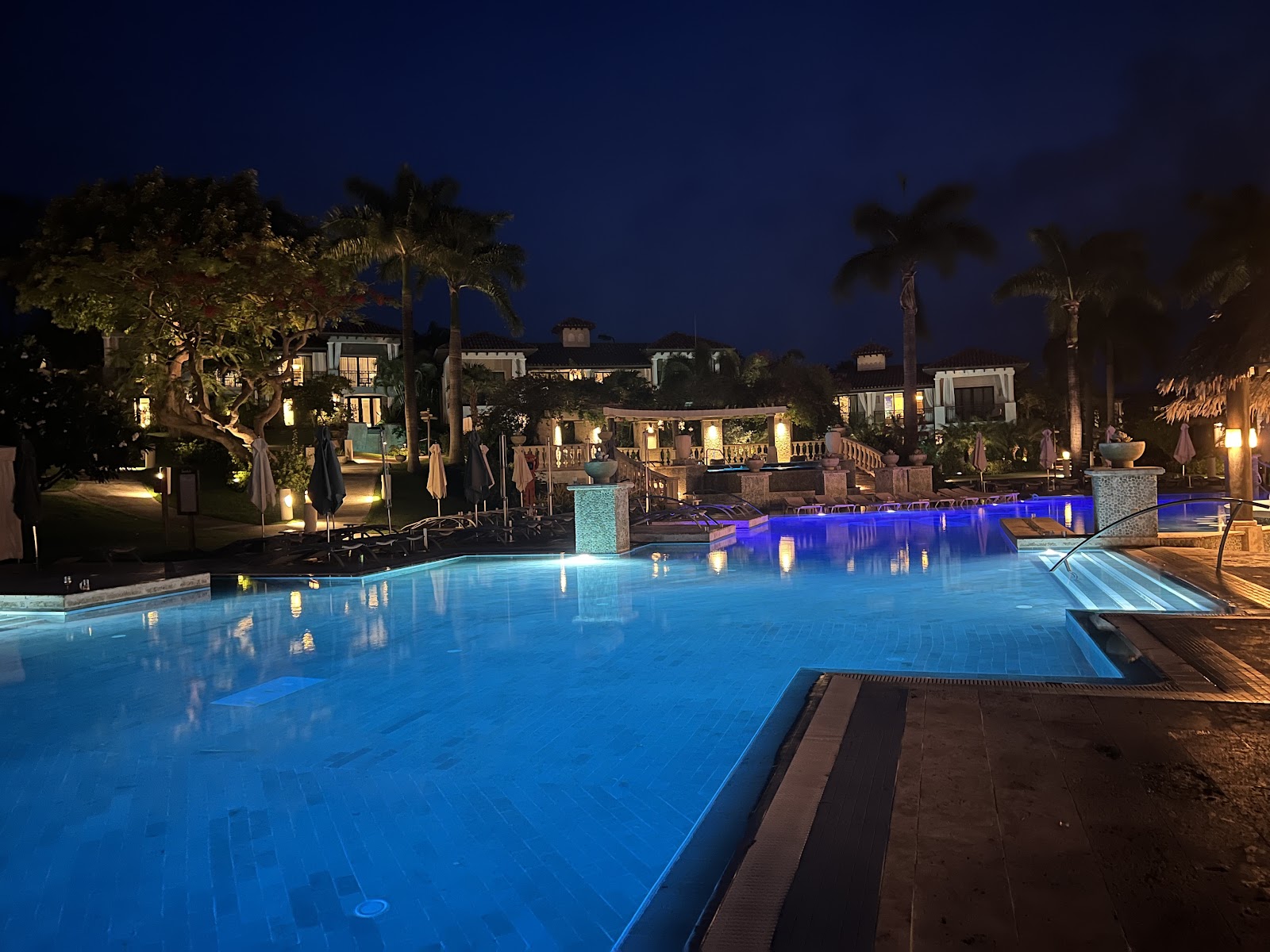 Illuminated pool complex at dusk with blue-lit water, surrounding colonial-style buildings, palm trees, and manicured grounds at Sandals Grenada.