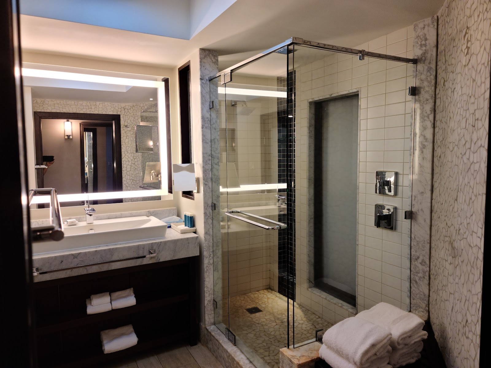 Modern bathroom with illuminated vanity mirror, marble countertop, glass-enclosed shower with black tile accent, and folded towels on floor.
