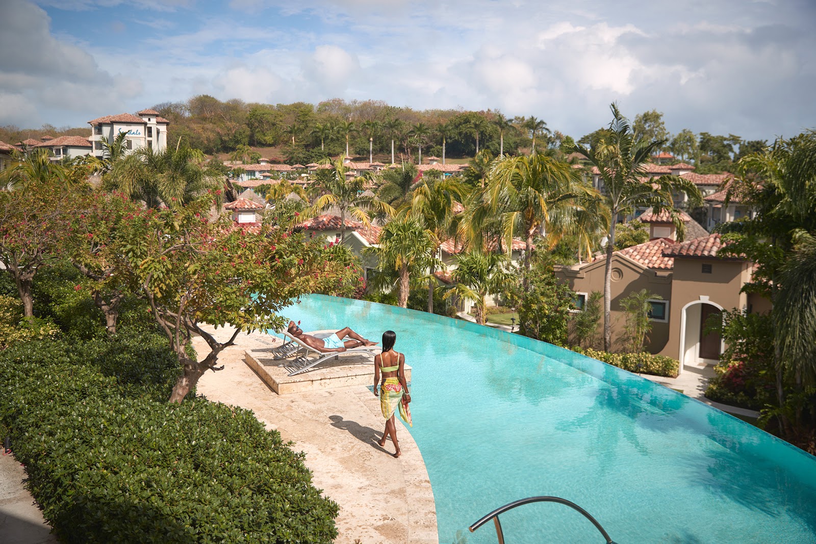 A woman in a swimsuit stands on the sand beside a turquoise infinity pool overlooking terracotta-roofed villas and palm trees at Sandals Grenada Resort & Spa.