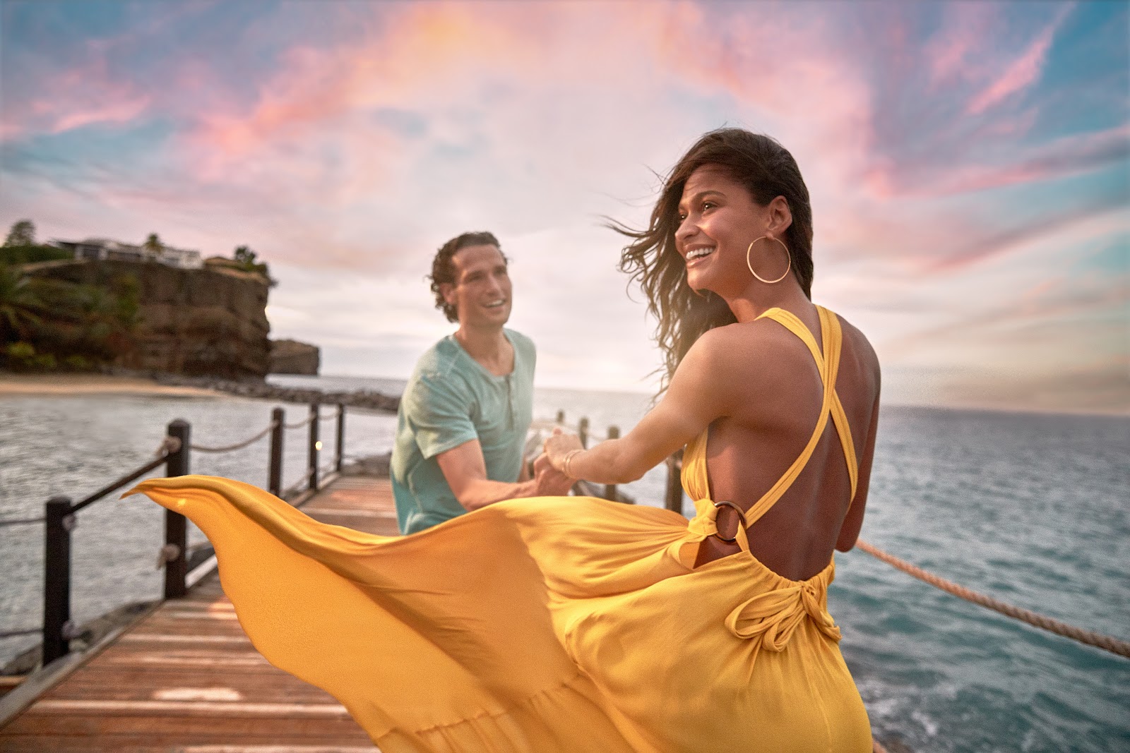 Woman in yellow dress twirls on wooden dock at sunset while man in turquoise shirt watches, with rocky coastline and ocean beyond.
