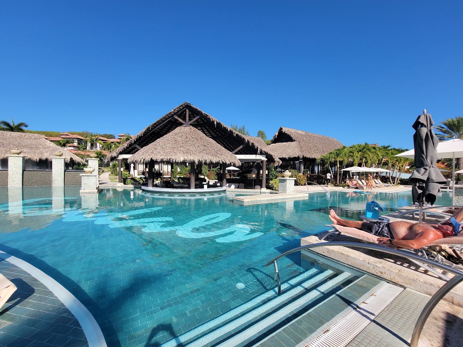 Turquoise pool with thatched-roof bar pavilion, palm trees, and guests lounging under umbrellas at Sandals Grenada Resort & Spa.
