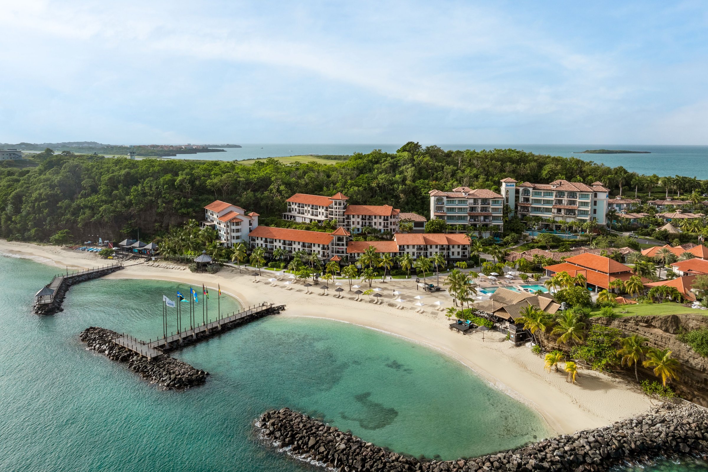 Aerial of the curved beach, breakwater lagoon, piers and terracotta-roofed resort.