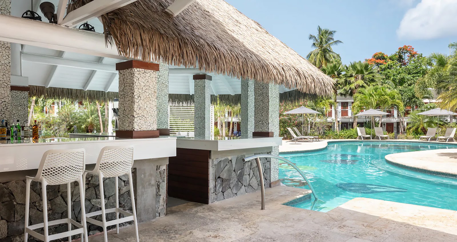 Beachfront bar with thatched roof, white counter seating, and turquoise pool beyond at Sandals Halcyon Beach, surrounded by palm trees.