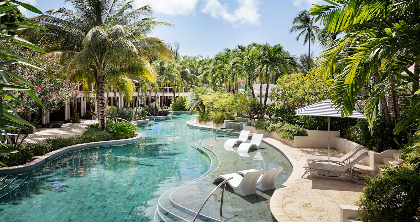 Turquoise lagoon-style pool winds through lush tropical grounds with lounge chairs, palm trees, and colonial-style buildings at Sandals Halcyon Beach.