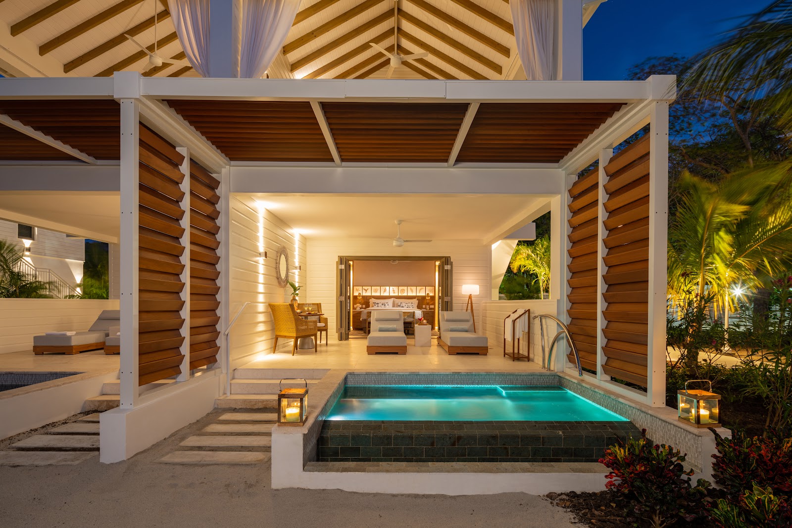 A private plunge pool illuminated at dusk beside an open-air suite with louvered walls and furnished patio at Sandals Halcyon Beach.