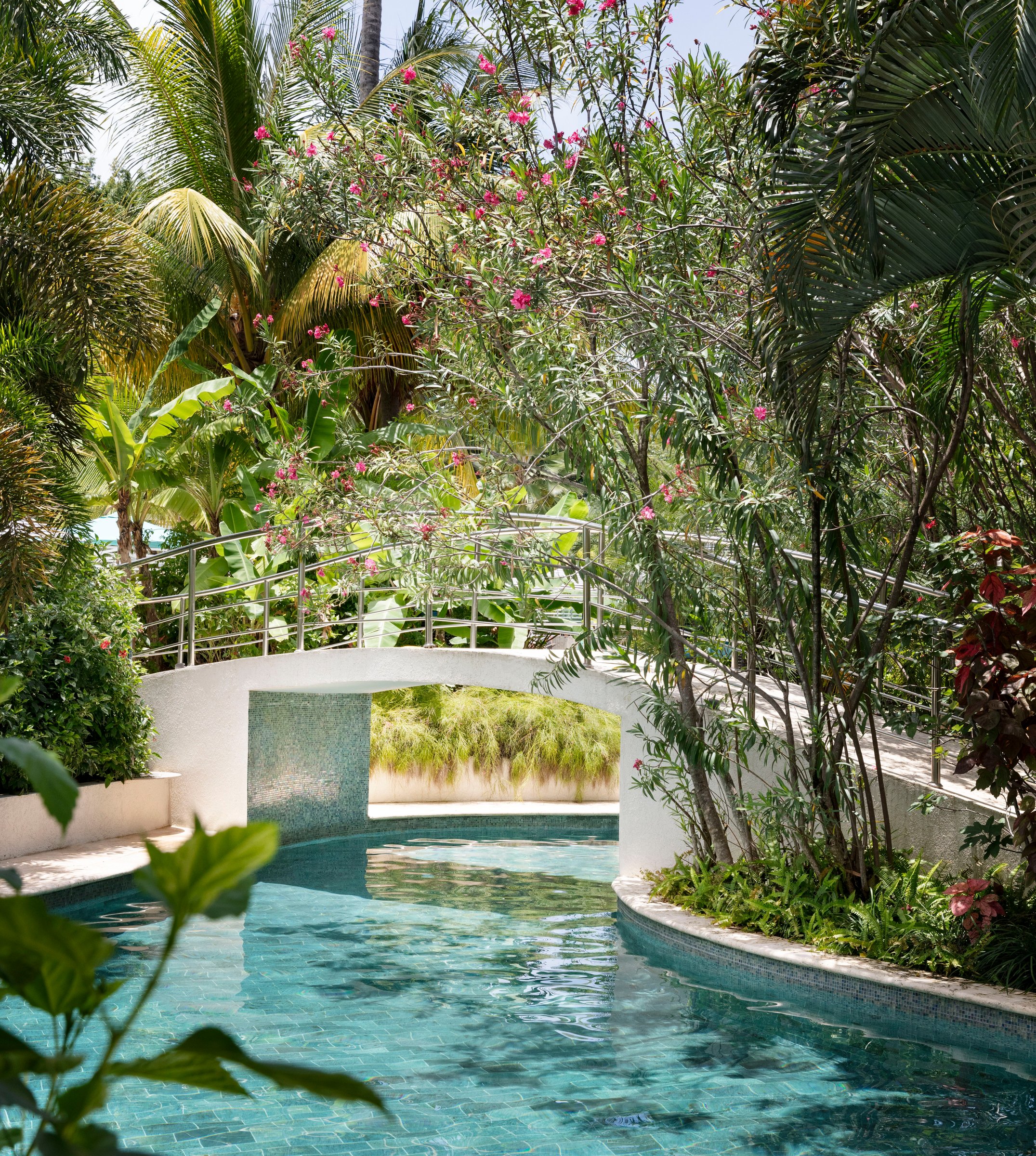 Lazy-river pool flowing under a white footbridge through flowering gardens.