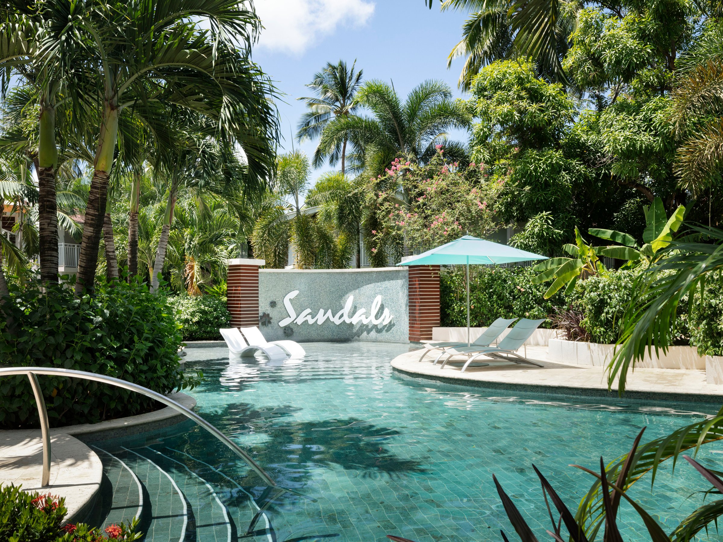 Lazy pool with in-water loungers beside a Sandals sign wall and umbrella.
