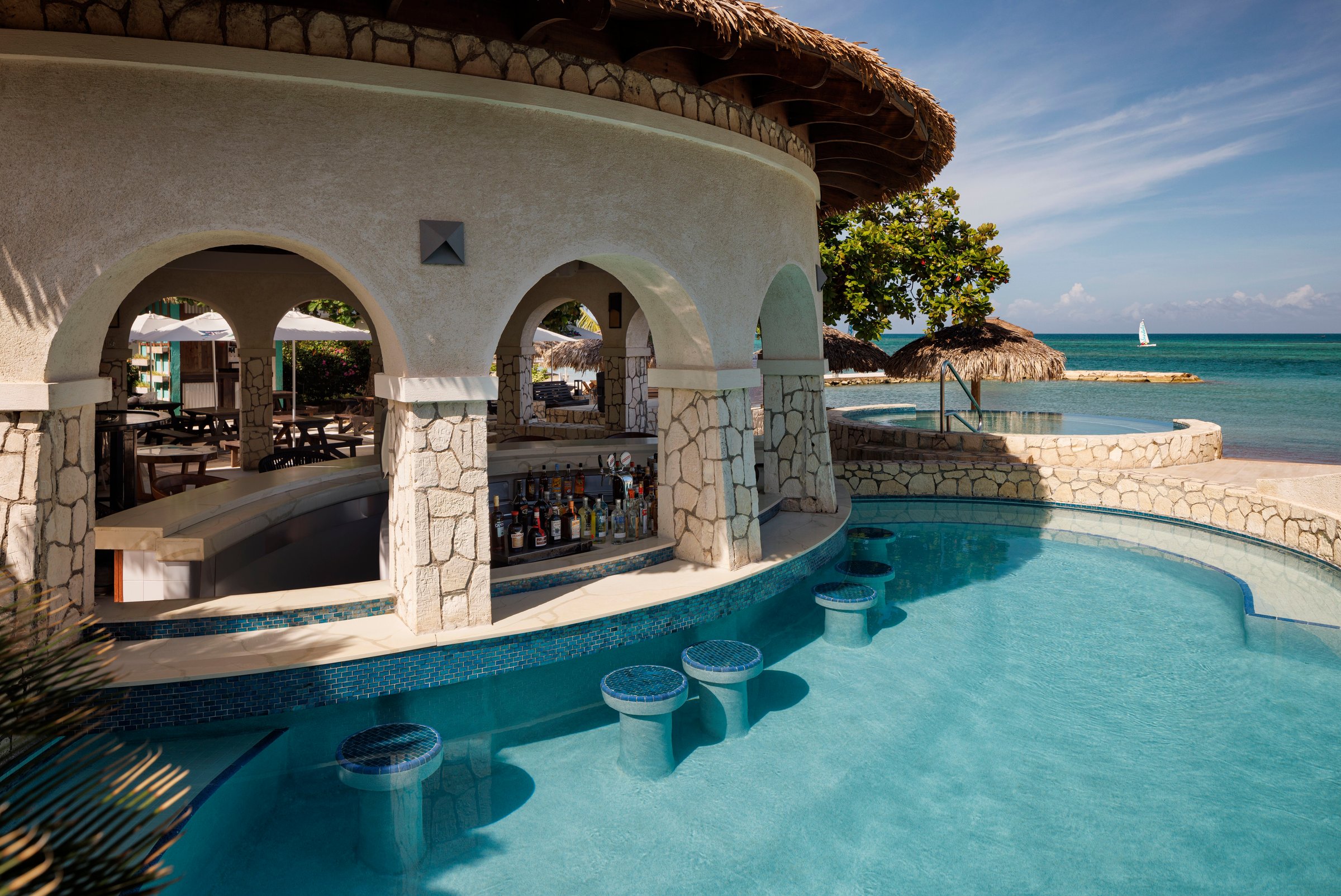 Swim-up bar under stone arches with in-pool stools and the sea beyond.