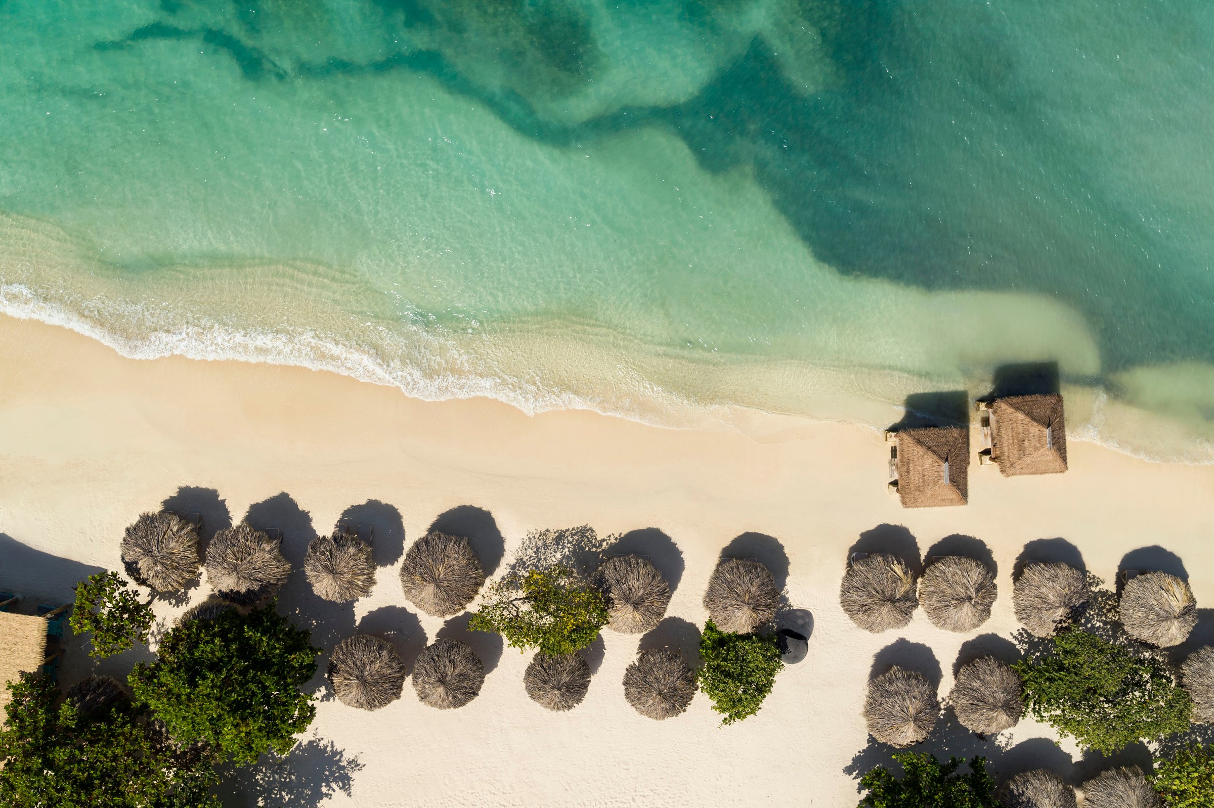 Top-down aerial of thatched umbrellas and cabanas on the white sand at the water line.