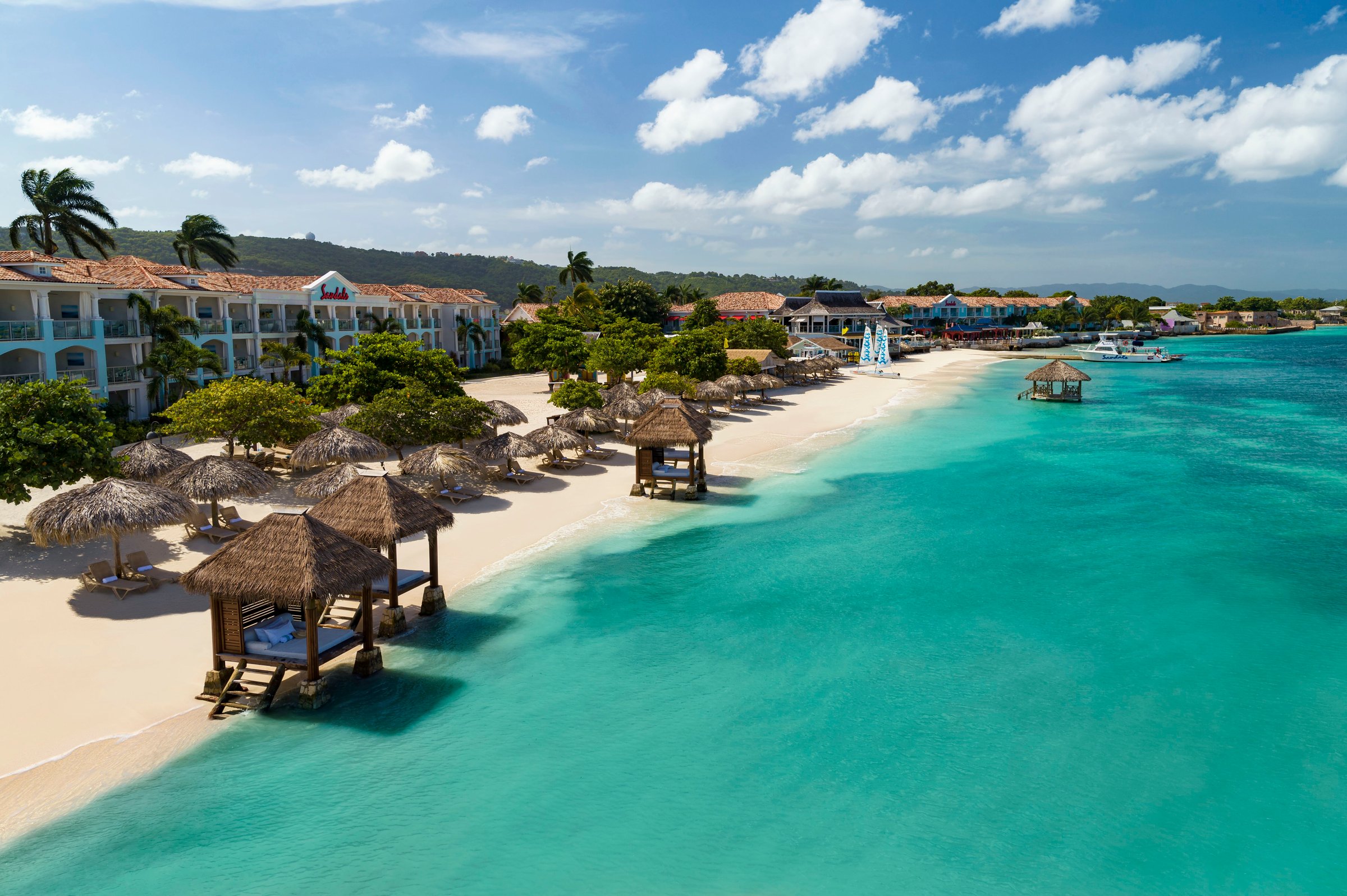 Low aerial of the beach with cabanas, an overwater gazebo and the resort buildings.