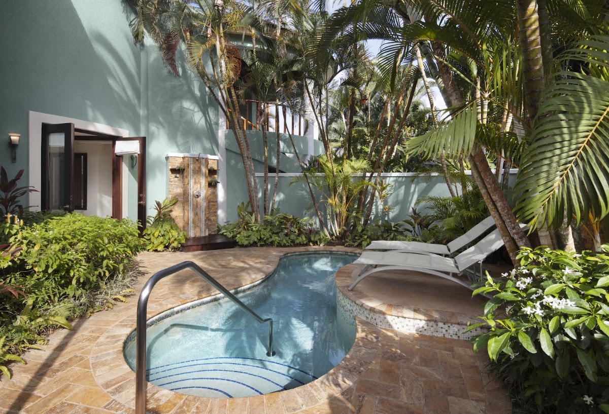 A small plunge pool with metal railing sits surrounded by tropical palms and lounge chairs beside a white villa with wooden doors at Sandals Negril.