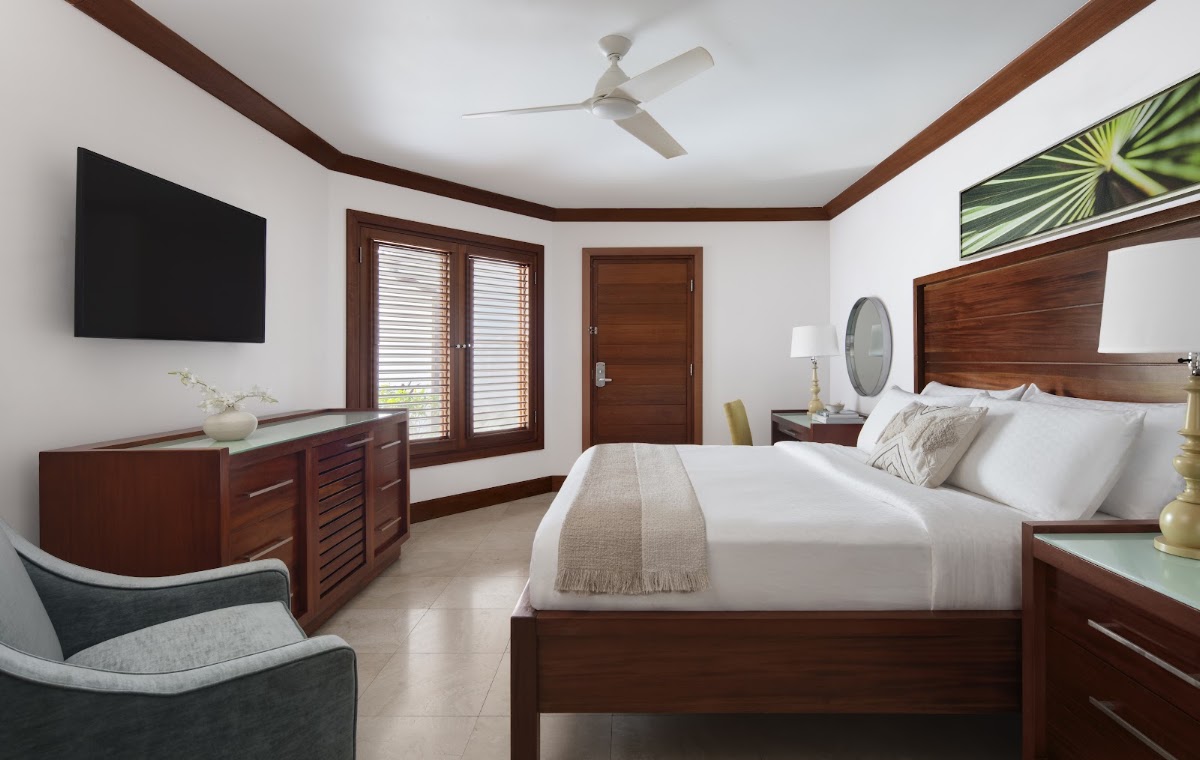 Spacious resort bedroom with white walls, dark wood furnishings, a king bed with white linens, ceiling fan, TV, and louvered doors opening to outdoor views.