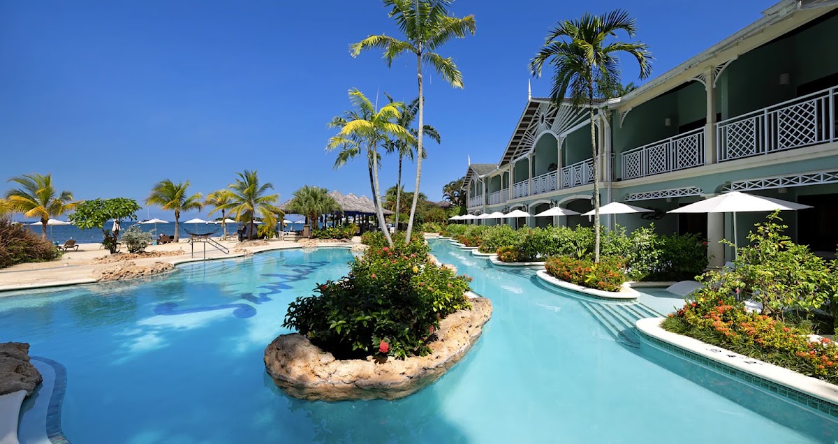 Curving lagoon-style pool with turquoise water winds past a green colonial-style building with white railings at Sandals Negril, flanked by palm trees and beach umbrellas.