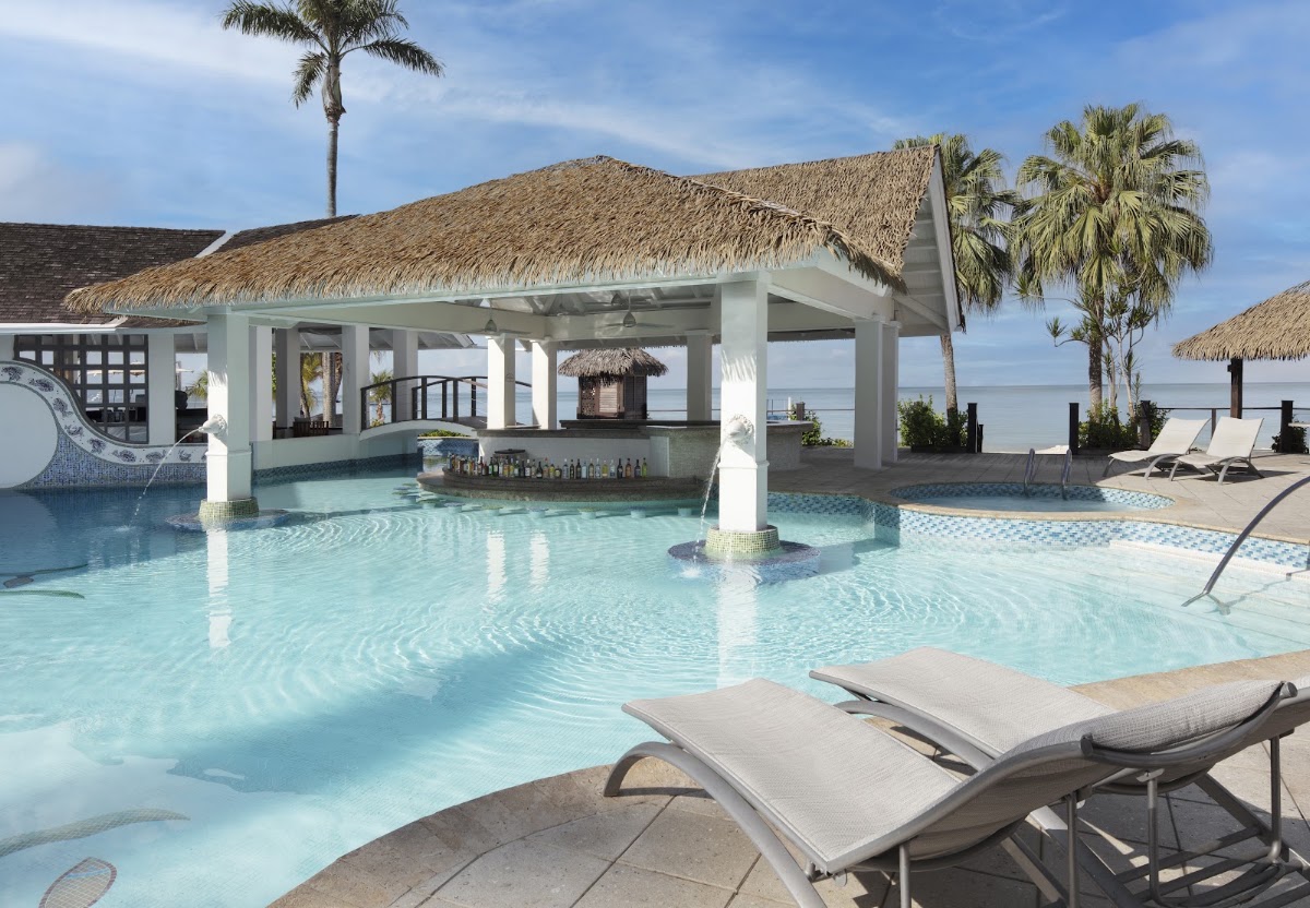Turquoise pool with white pergola bar, thatched-roof structures, palm trees, and ocean view at Sandals Negril.