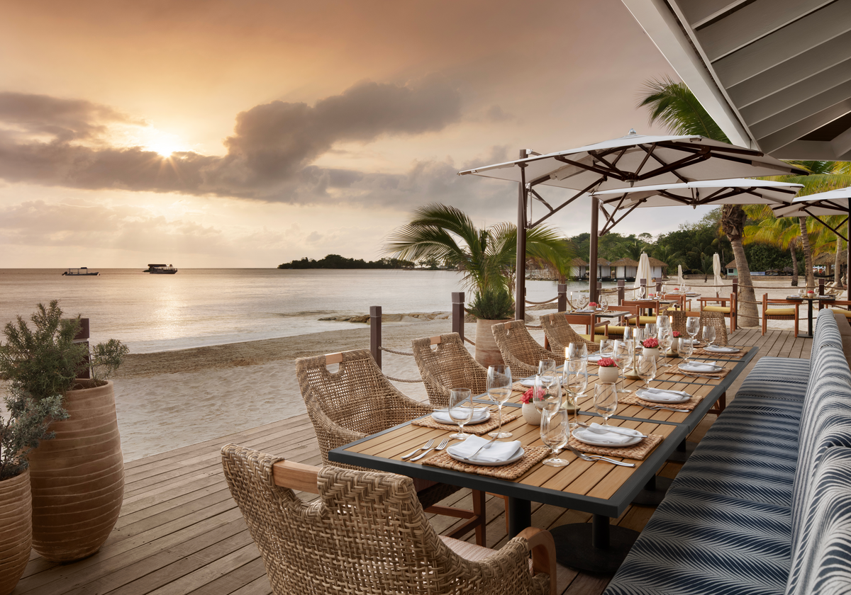 Long wooden deck dining table set for dinner with woven chairs and umbrellas overlooking calm water at Sandals Negril.