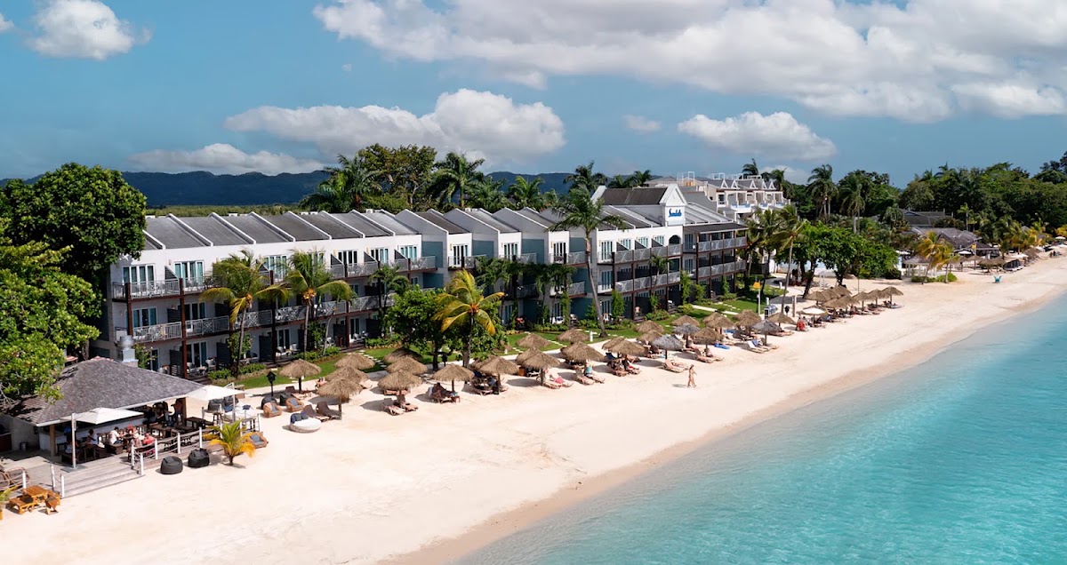 Sandals Negril beachfront with white colonial-style buildings, palm trees, sandy beach, and turquoise water under partly cloudy sky.