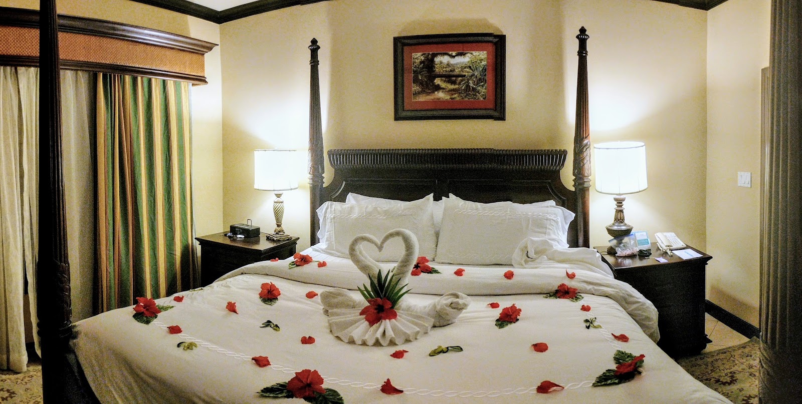 Four-poster bed decorated with red flower petals and towel art in a resort bedroom with cream walls and black furnishings.