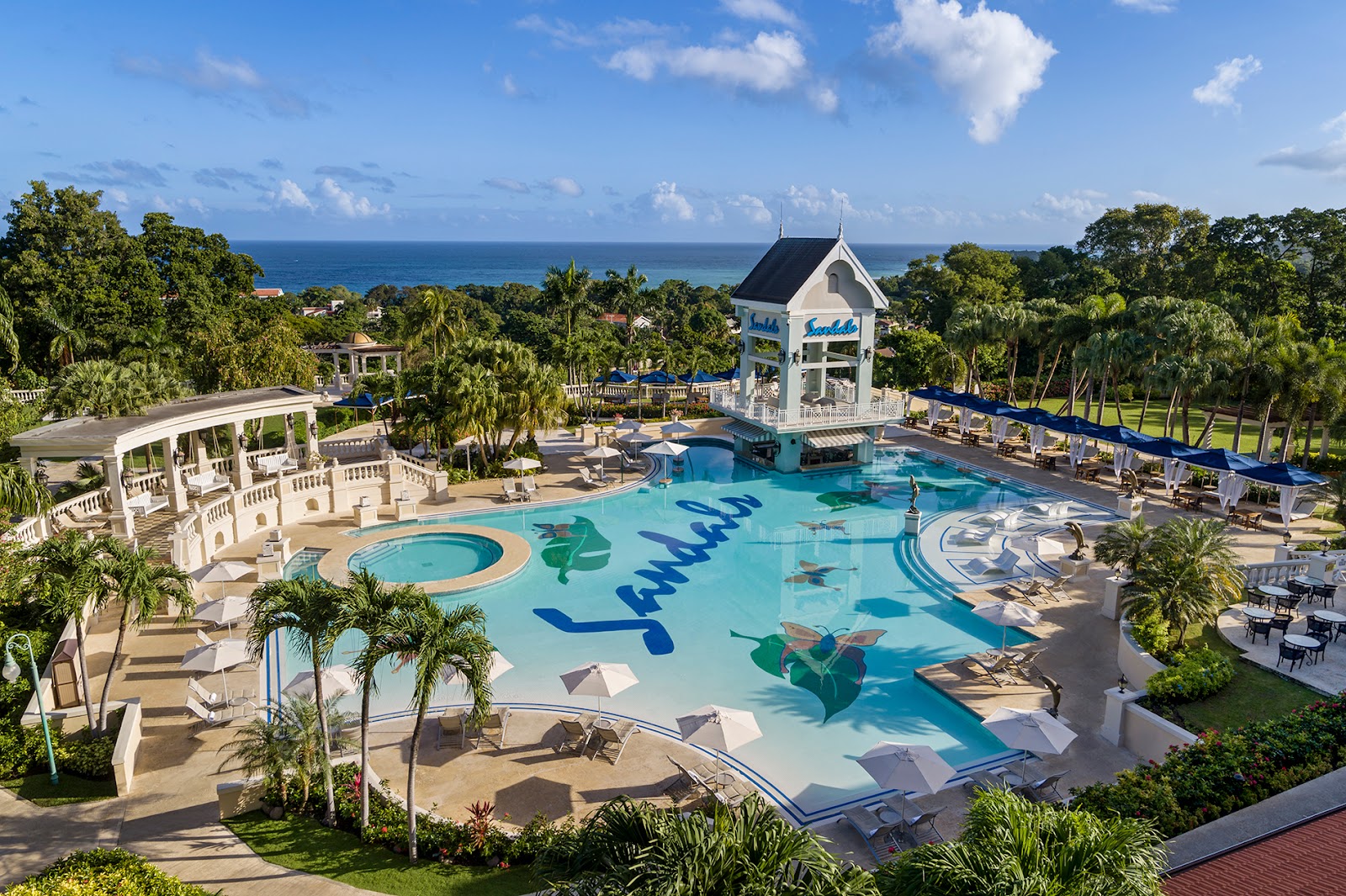 Sandals Ochi Beach Resort pool complex with turquoise water, white gazebo, palm trees, and ocean view beyond lush grounds.