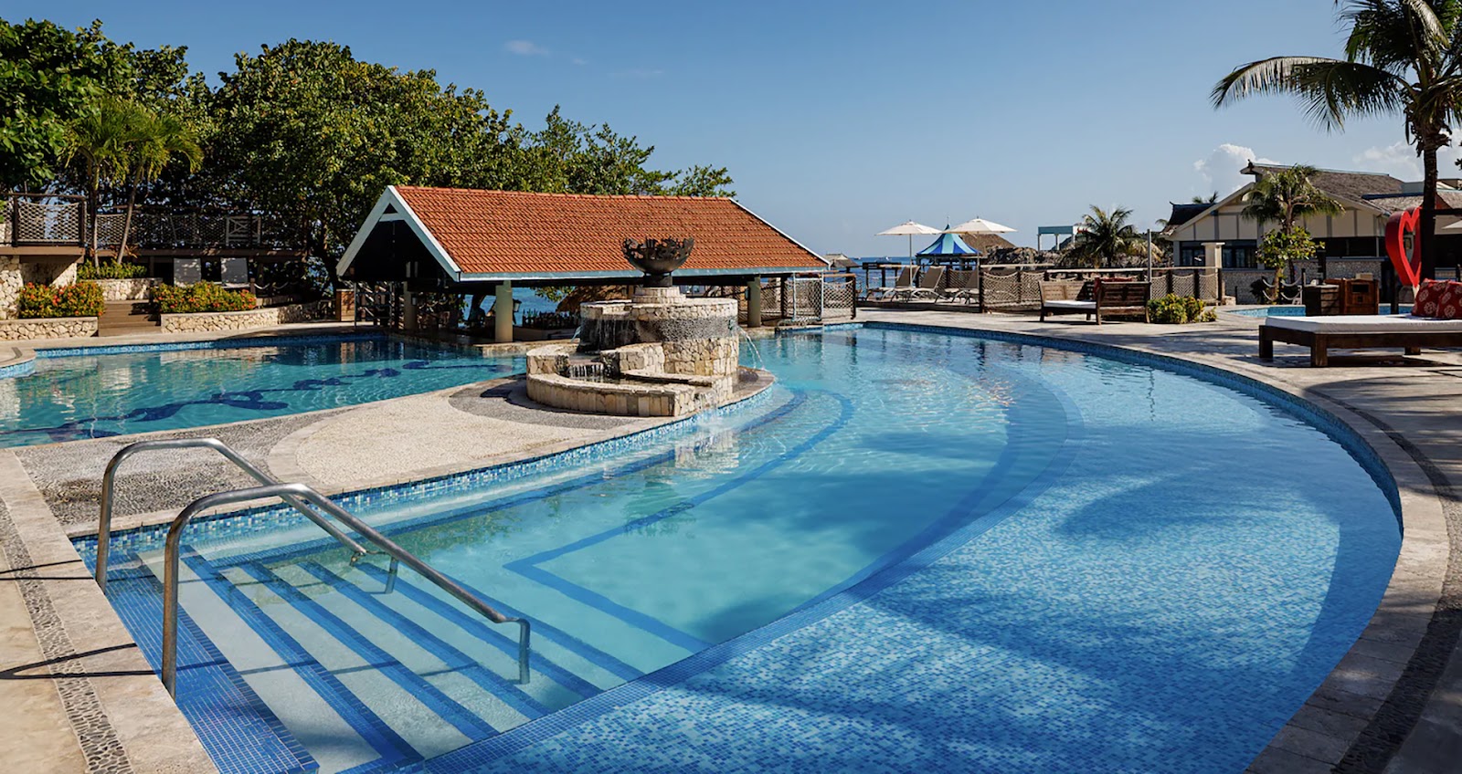 Curved swimming pool with shallow steps at Sandals Ochi Beach Resort, stone bar structure in center, thatched roof cabana beyond, palm trees and clear sky.