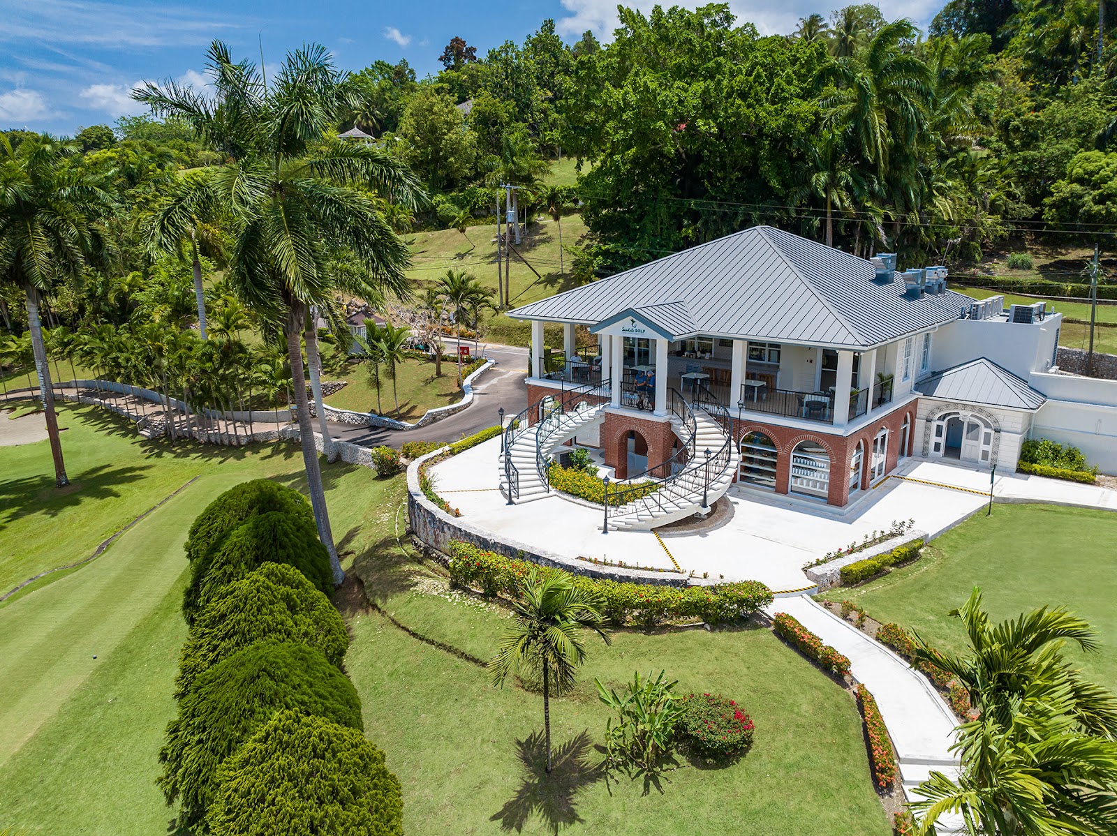 Aerial view of Sandals Ochi Beach Resort's white main building with metal roof surrounded by manicured lawns, palm trees, and lush tropical vegetation.