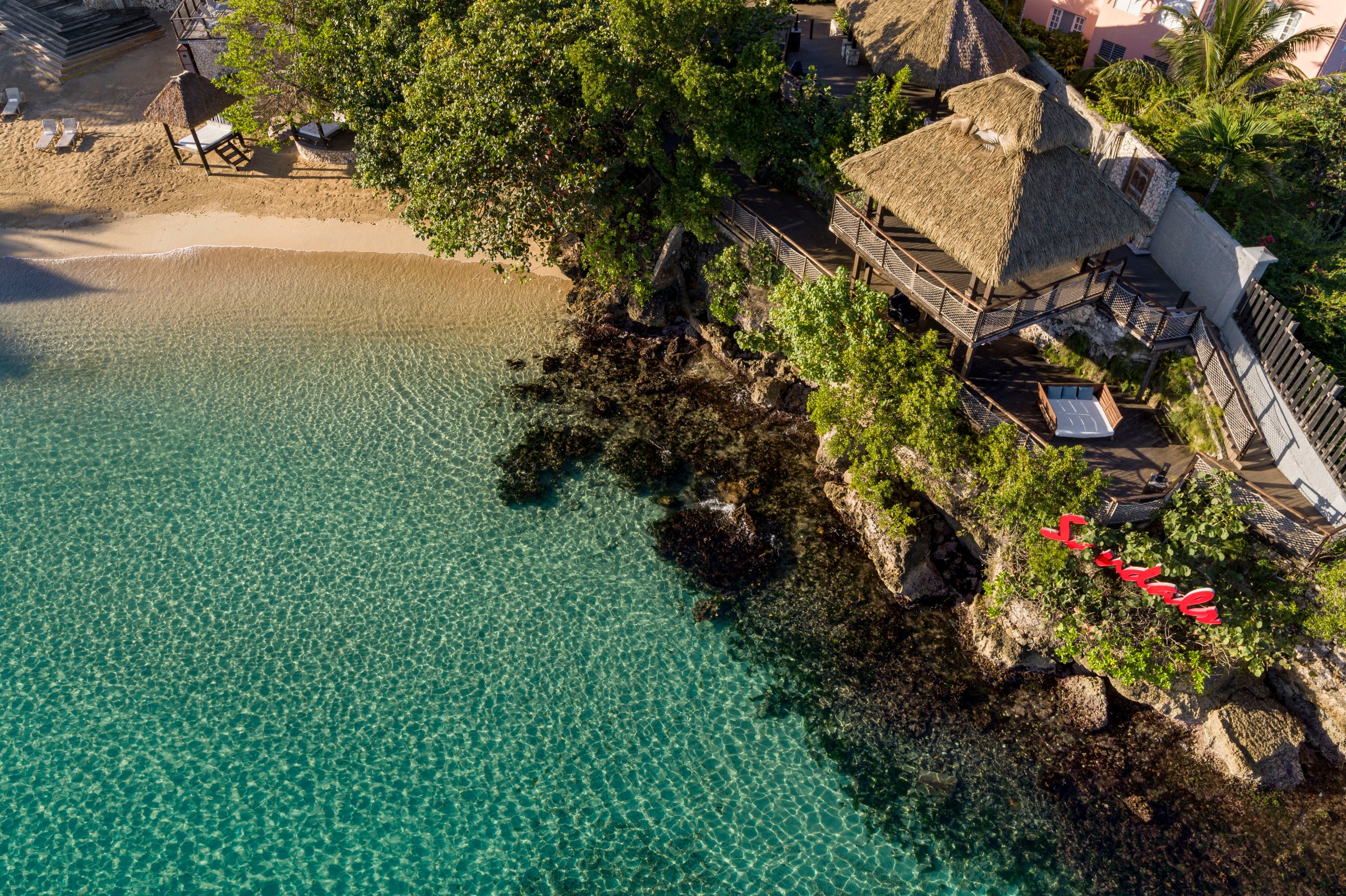Aerial of a clifftop thatched gazebo above a clear-water cove.