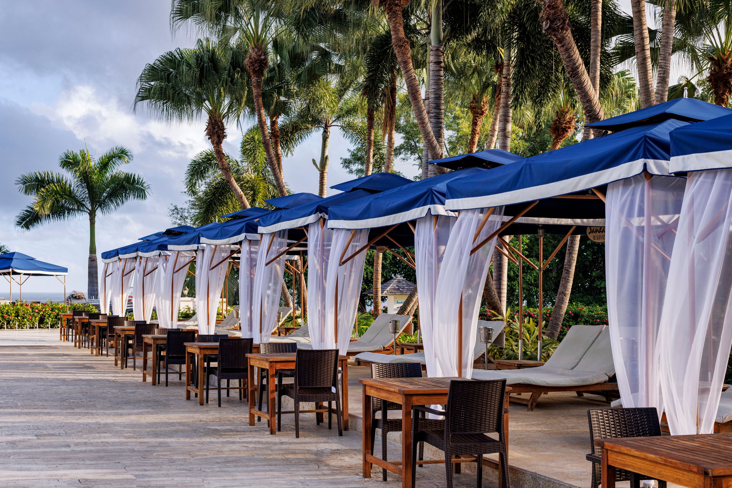 Row of blue-topped poolside cabanas with tables under palms.