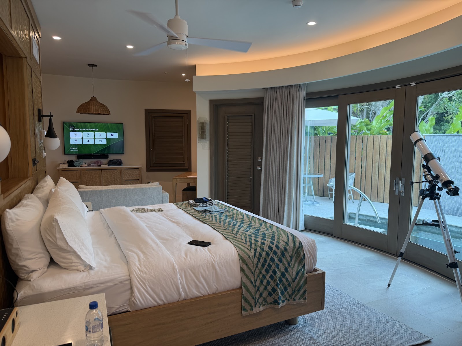 Resort bedroom with king bed, balcony overlooking tropical landscape and water, sitting area, and ceiling fan.