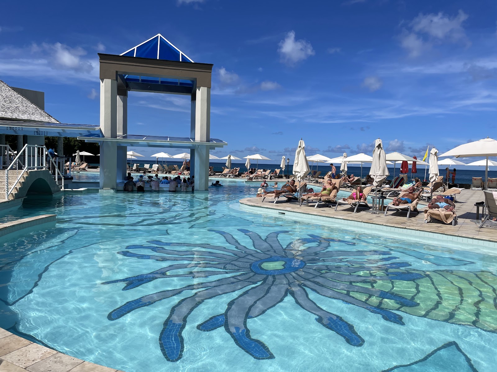 Resort pool with decorative octopus mosaic design, white umbrellas, and ocean view beyond at Sandals Regency La Toc.