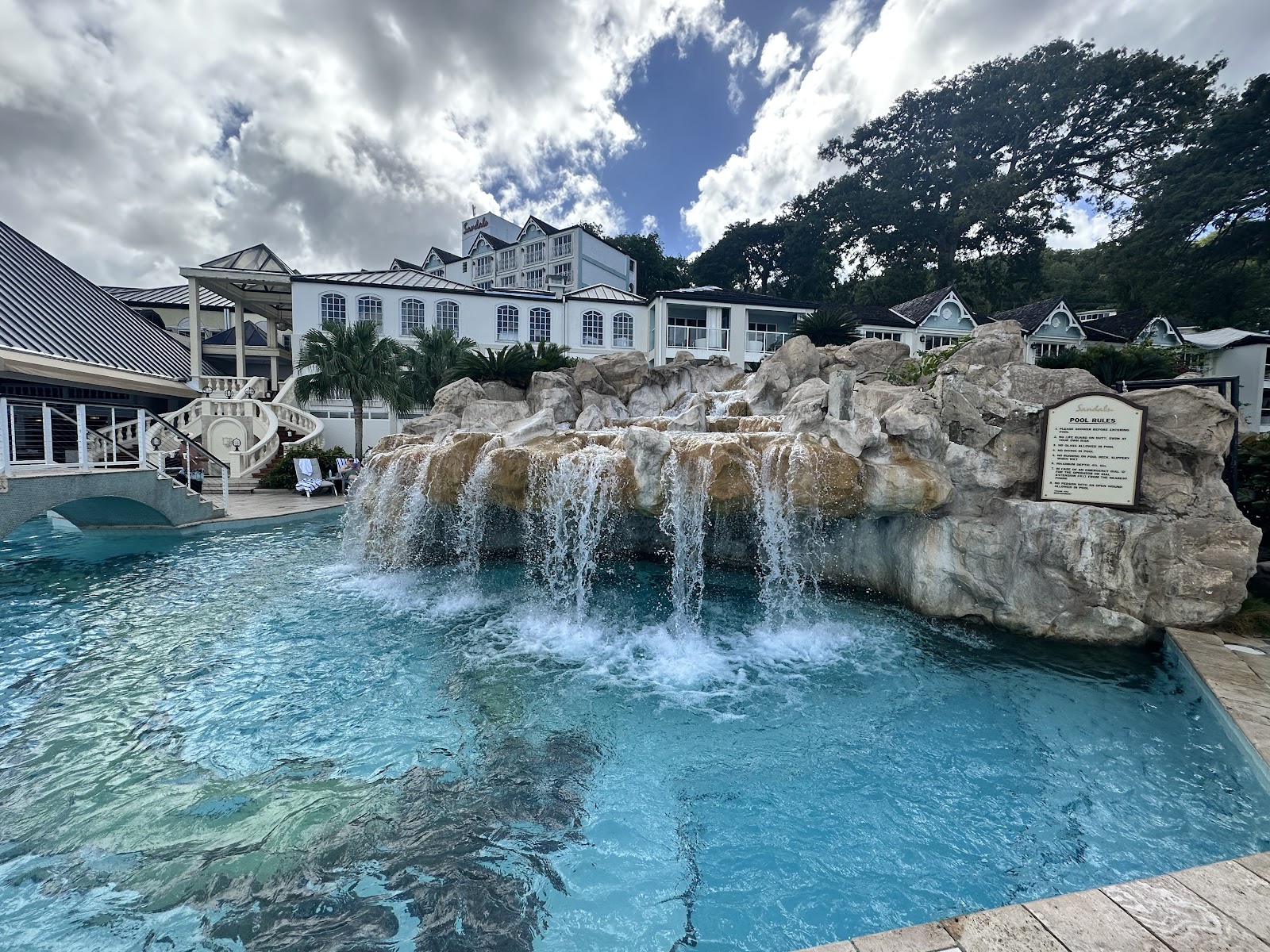 Pool with cascading waterfall over large rocks at Sandals Regency La Toc, white main building and palm trees in background.
