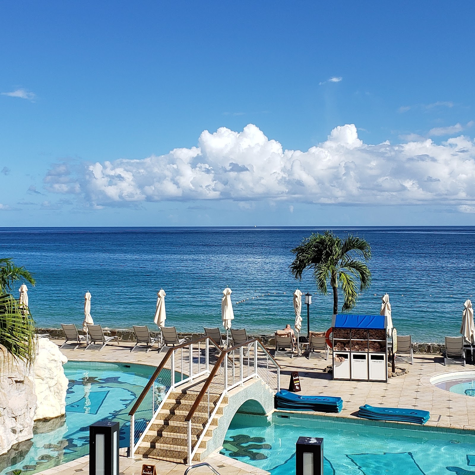 Resort pools and beach area with lounge chairs and white umbrellas overlooking turquoise Caribbean waters under a cloudy blue sky.