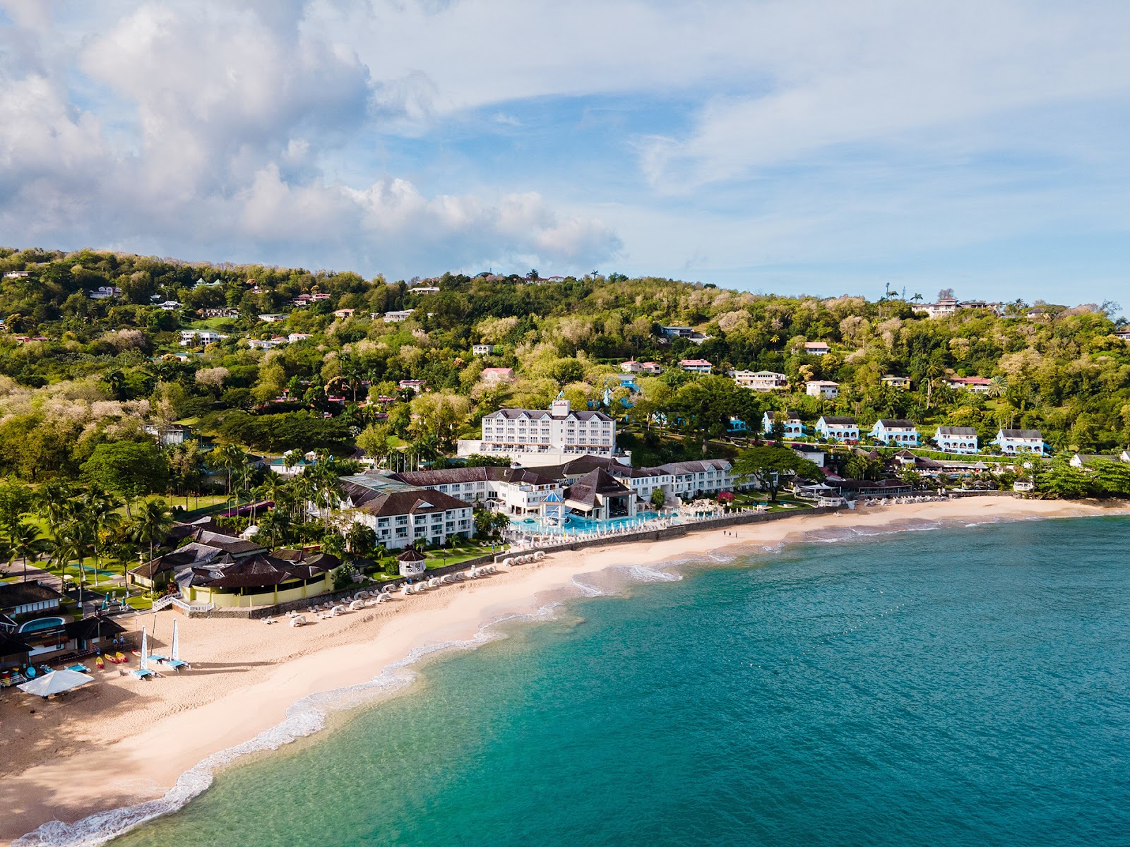 Sandals Regency La Toc resort sits on a sandy beach with turquoise water, surrounded by lush hillside vegetation and colonial-style buildings.