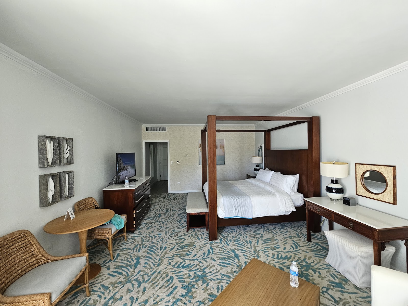 Spacious resort suite with wood four-poster bed, patterned carpet, sitting area with wicker chairs, and dark wood furnishings.