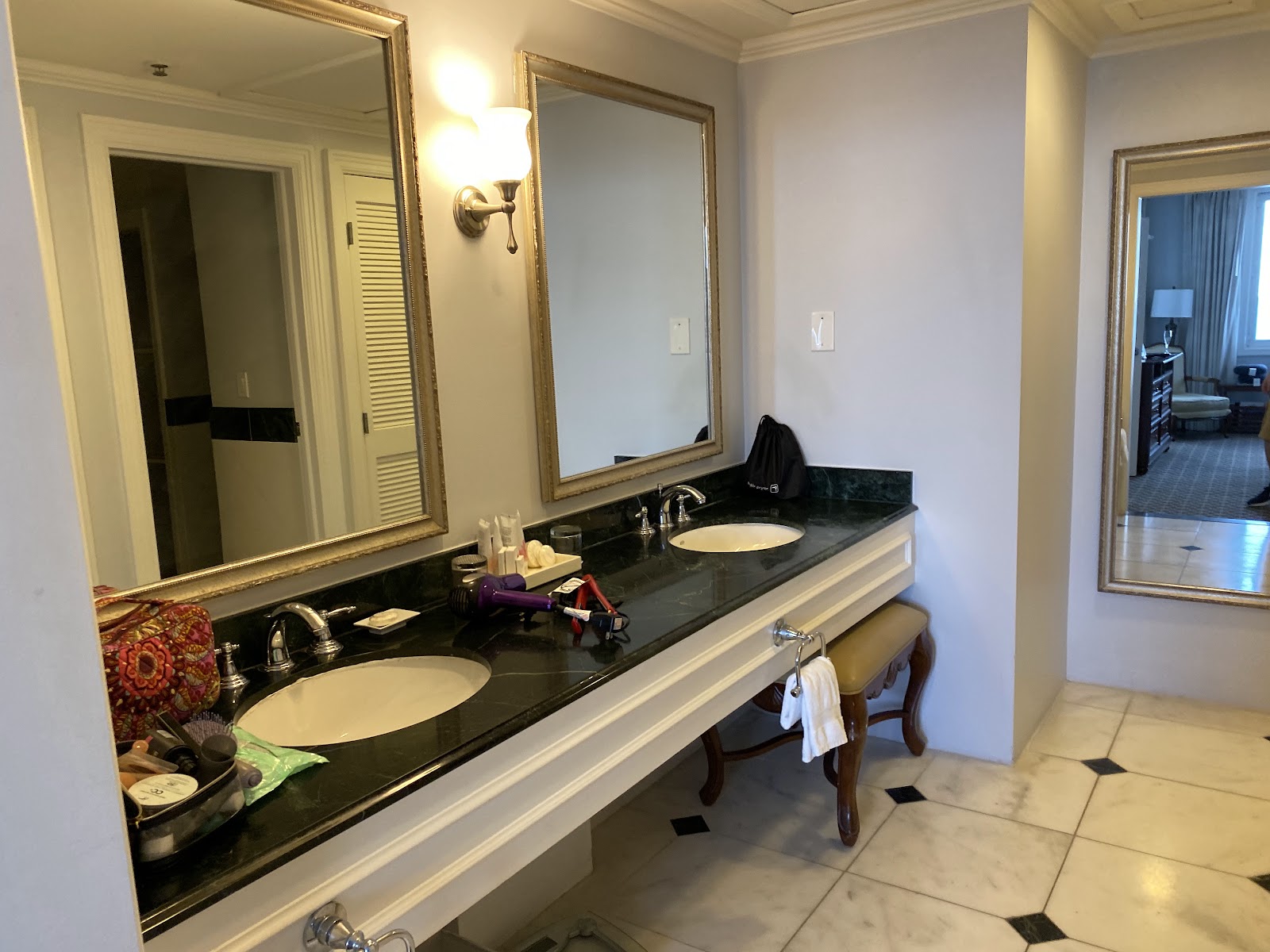 Resort bathroom vanity with dual sinks, black countertop, gold-framed mirrors, and cream-colored cabinetry with decorative bench seating.