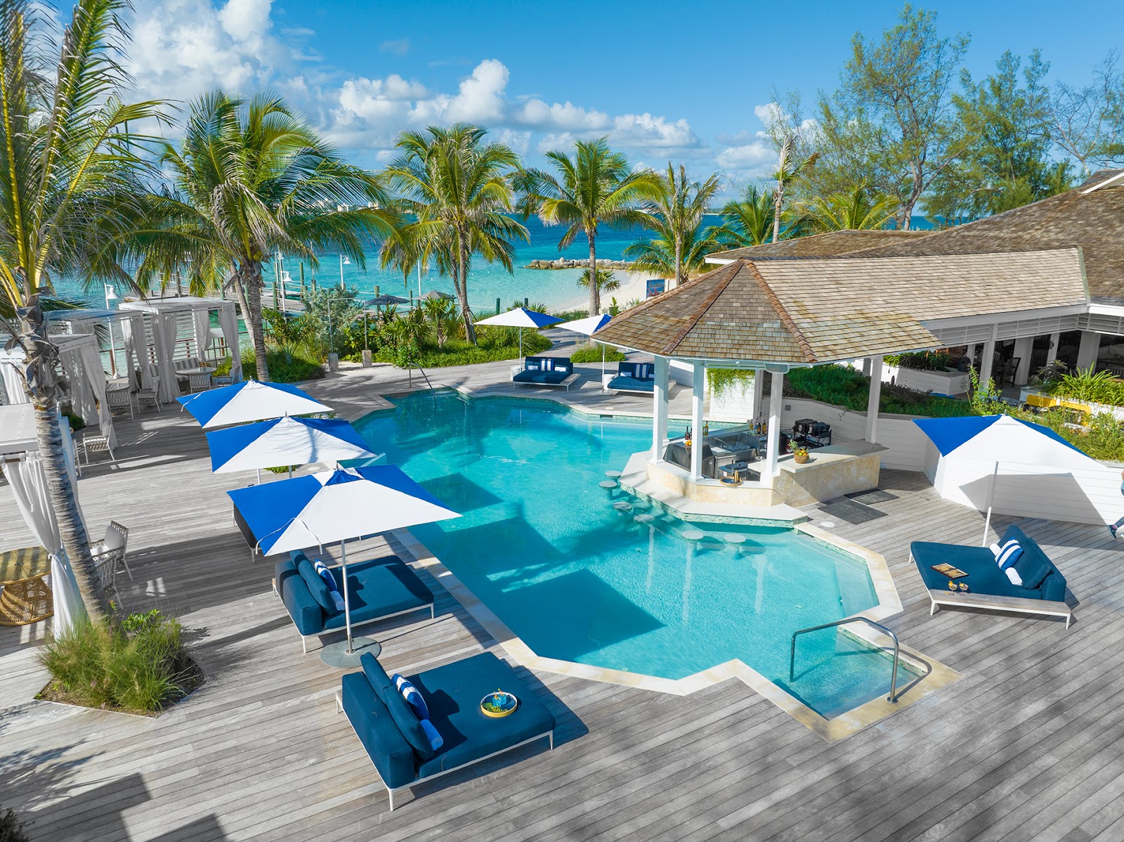 Pool deck at Sandals Royal Bahamian with blue and white umbrellas, lounge chairs, gazebo, and turquoise ocean visible beyond palm trees.