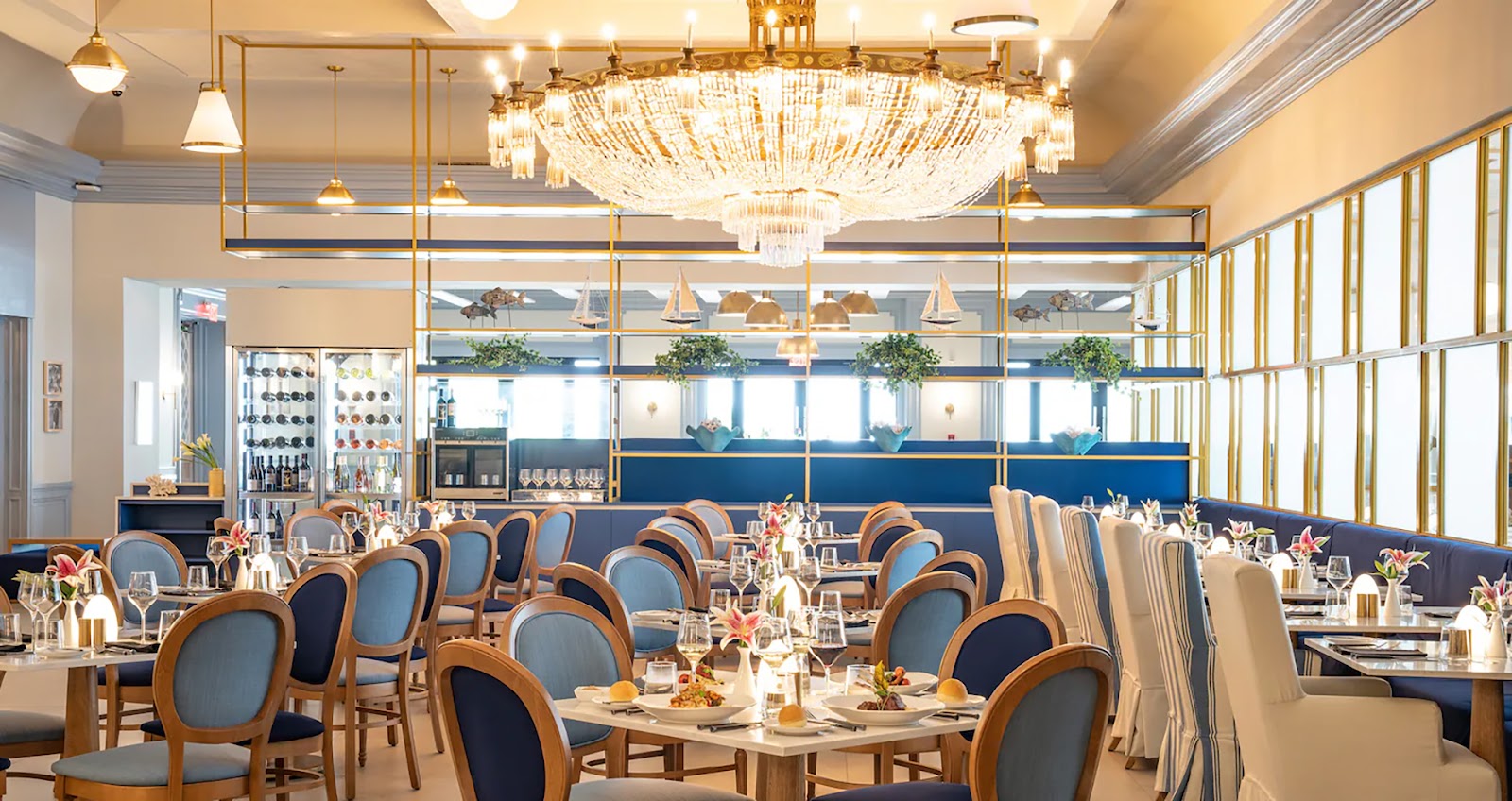Elegant dining room with wooden chairs upholstered in blue, round tables set for service, crystal chandelier overhead, and large windows overlooking the ocean.