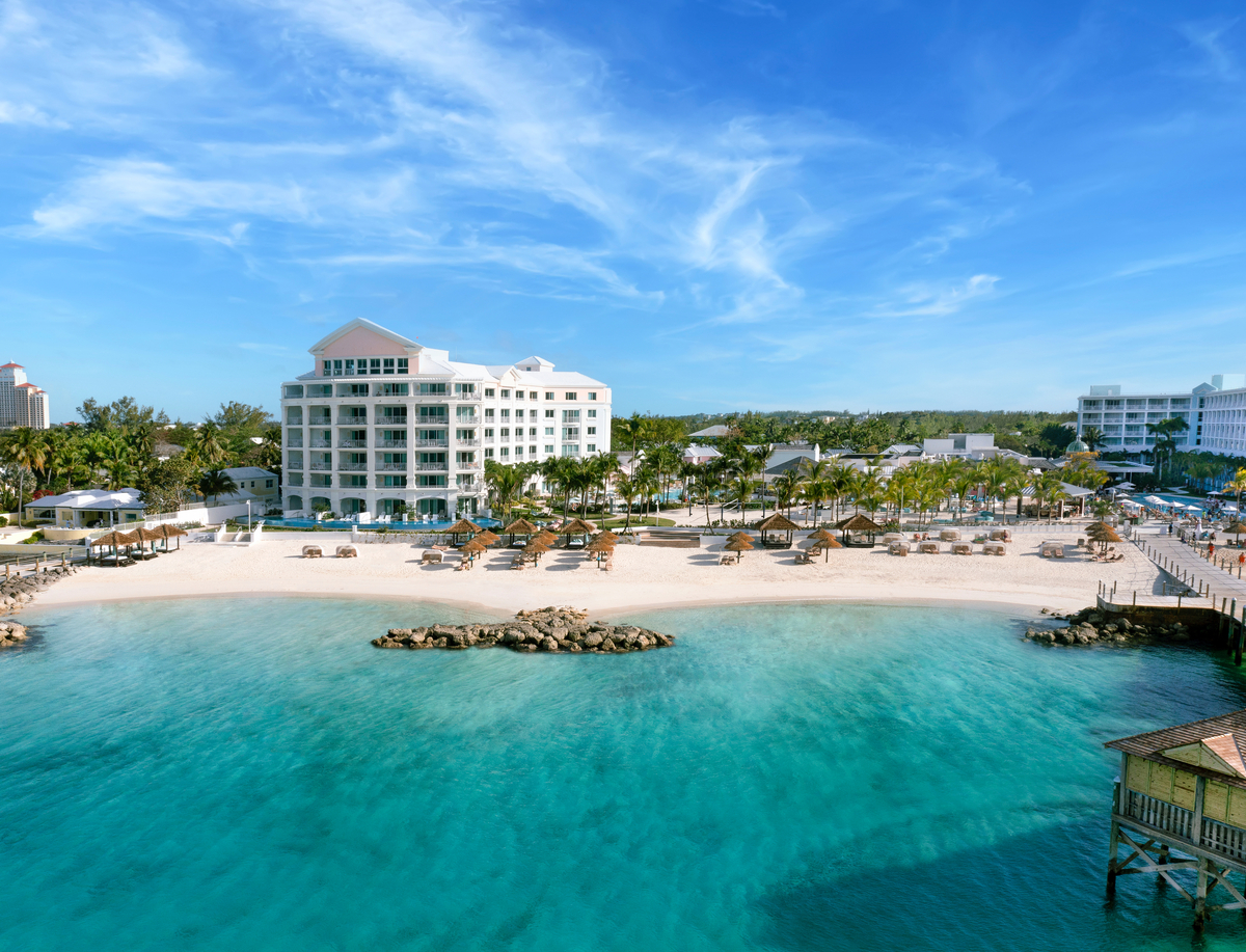 White multi-story resort building with turquoise ocean, sandy beach with lounge chairs, palm trees, and wooden pier under blue sky.