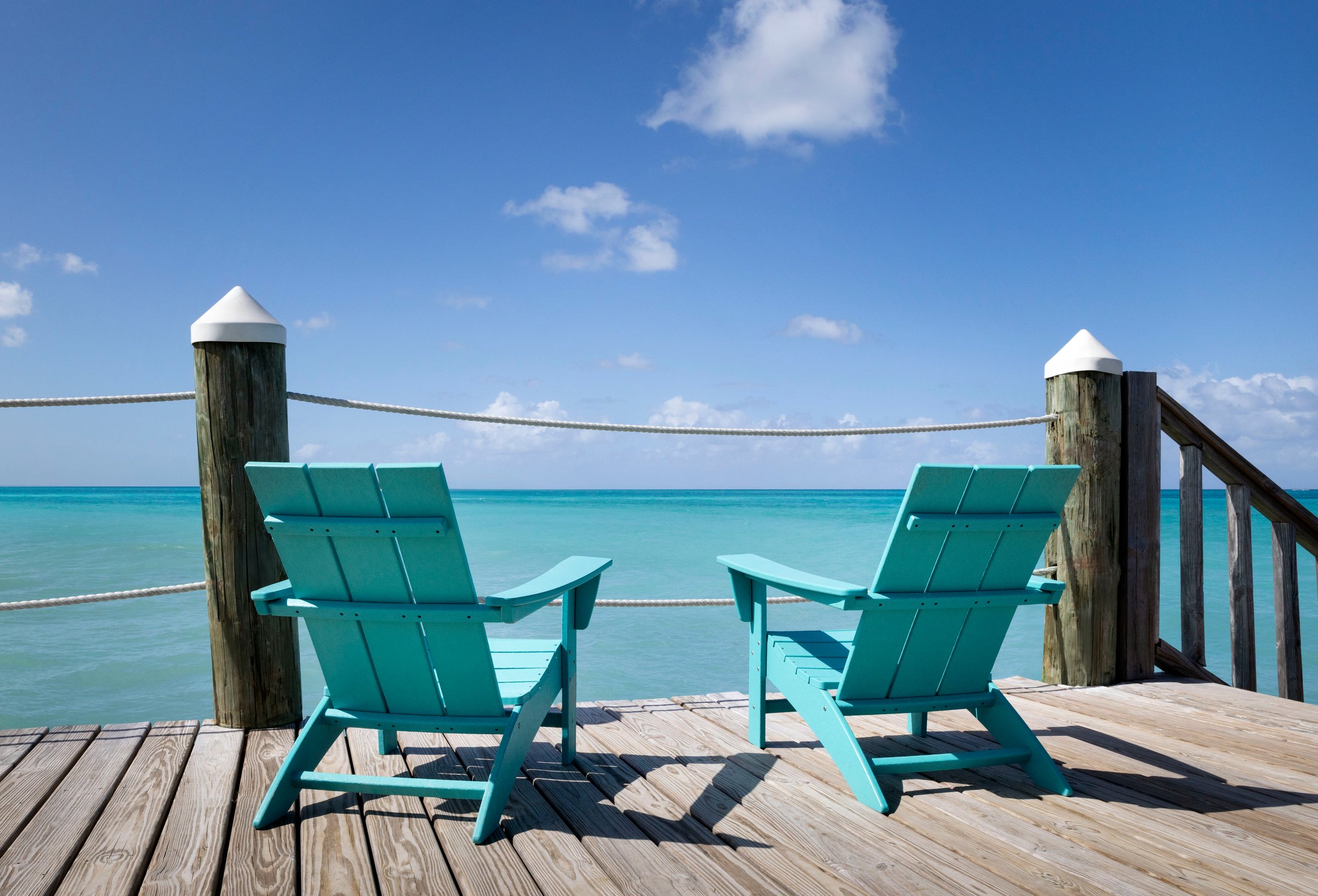 Two teal Adirondack chairs on a wooden pier facing the sea.