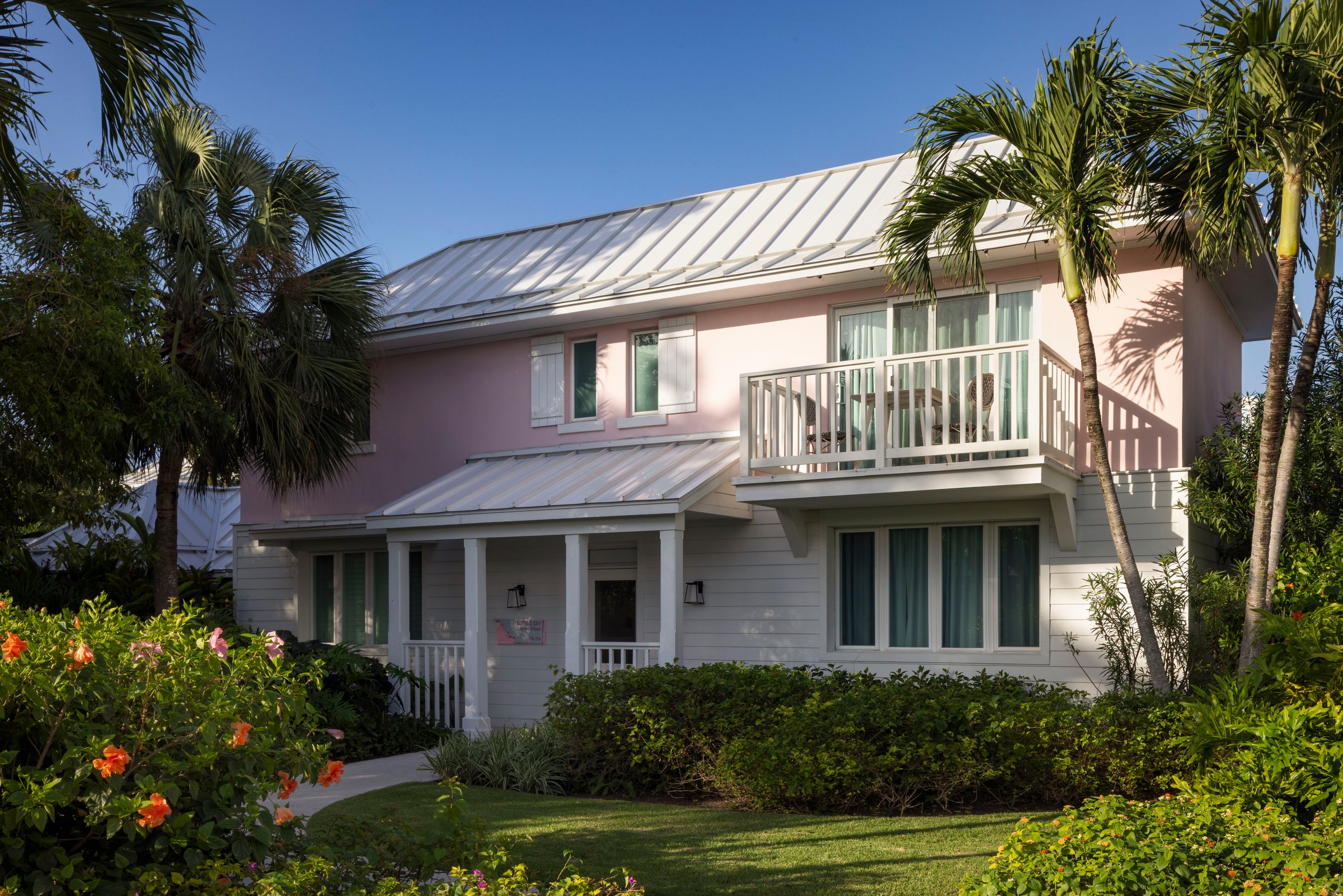 Pink two-storey villa with a balcony among palms and flowering shrubs.