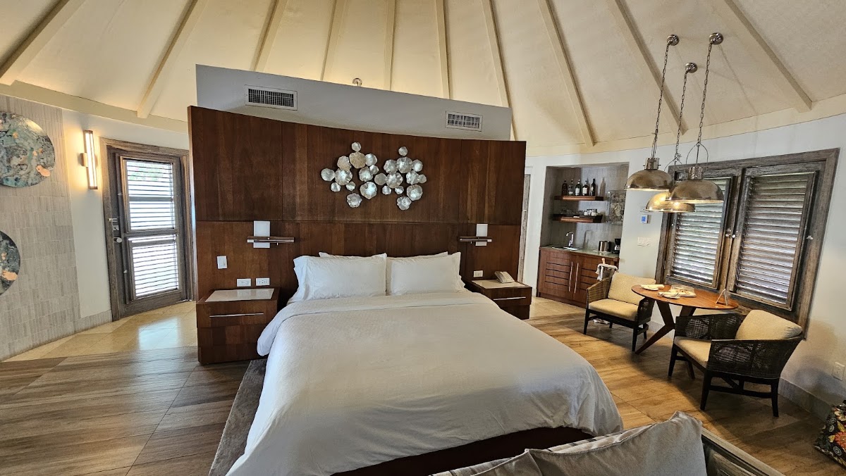 Spacious overwater villa bedroom with vaulted ceiling, dark wood bed frame, white linens, decorative plate wall art, and lounge seating near glass doors.