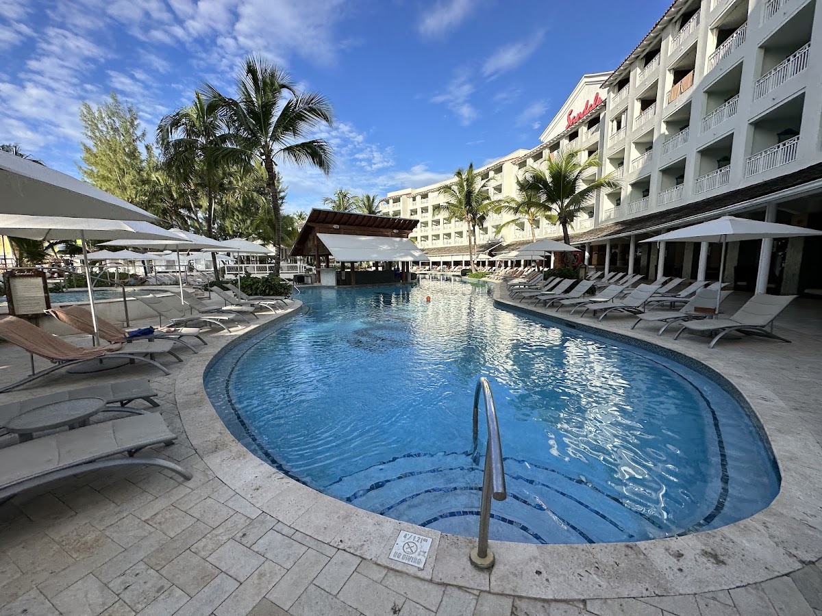 Curved blue pool with lane markers surrounded by lounge chairs and white umbrellas, flanked by multi-story resort buildings and palm trees under clear sky.