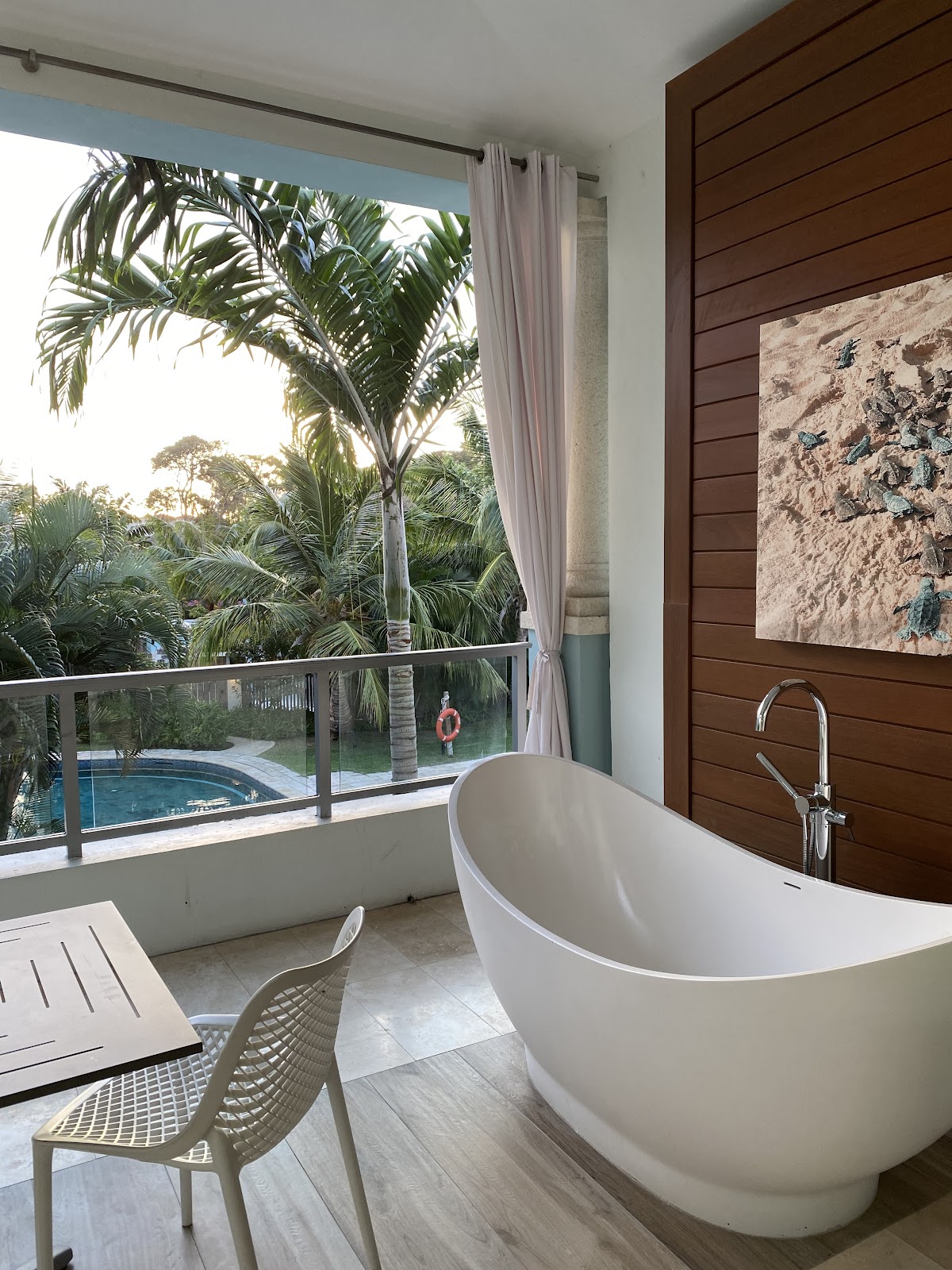 Bathroom with freestanding white tub, dark wood wall paneling, and floor-to-ceiling window overlooking palm trees and resort pool below.