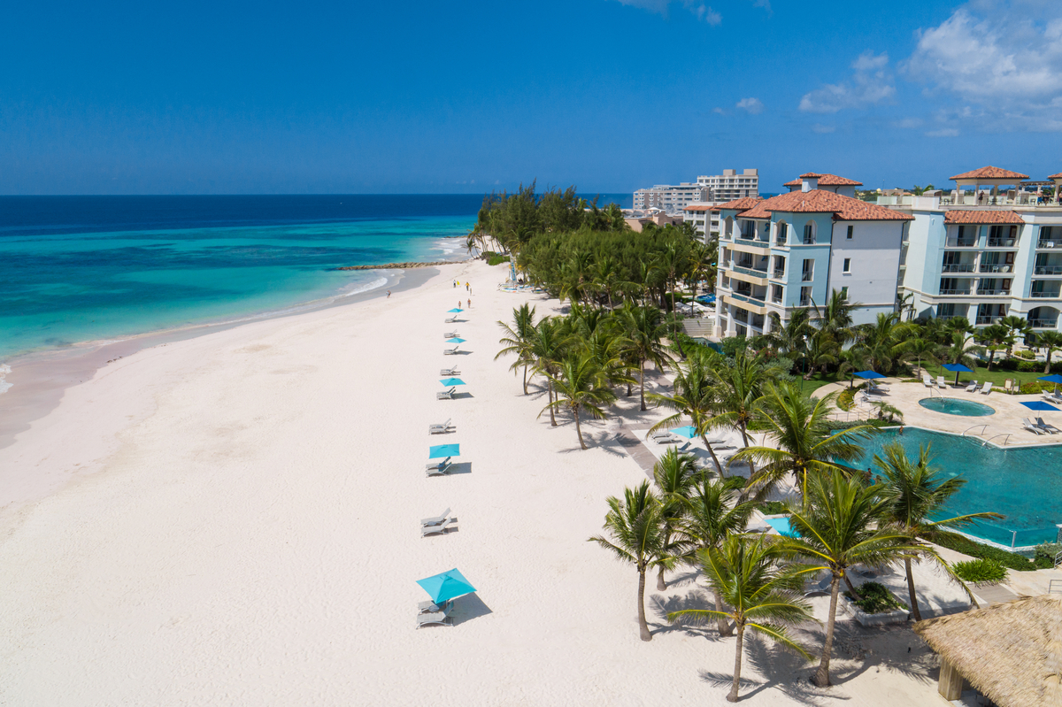 White sand beach with blue umbrellas and loungers, turquoise ocean, and Sandals Royal Barbados resort buildings with red-tile roofs and pool area in background.