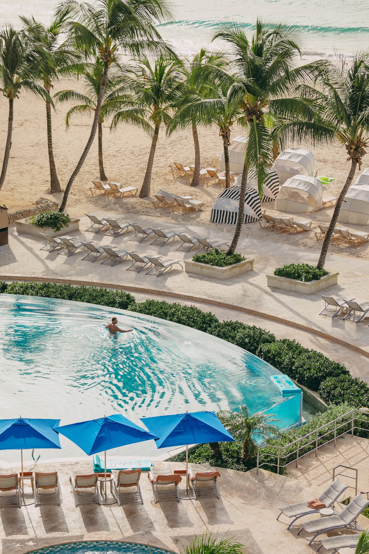 Resort pool with blue umbrellas and lounge chairs overlooks sandy beach with palm trees and turquoise ocean beyond.
