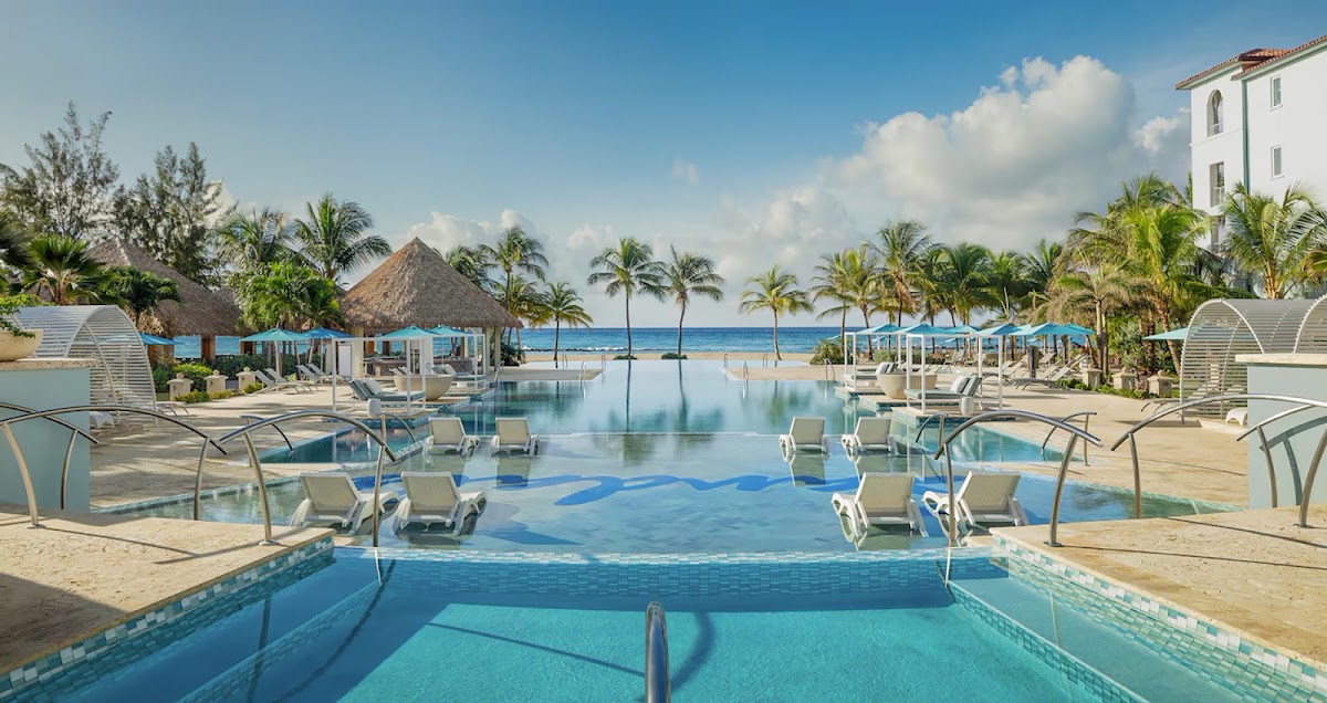 Swim-up bar in foreground with tiered pools, lounge chairs, and thatch-roof cabanas leading to the ocean and palm trees at Sandals Royal Barbados.