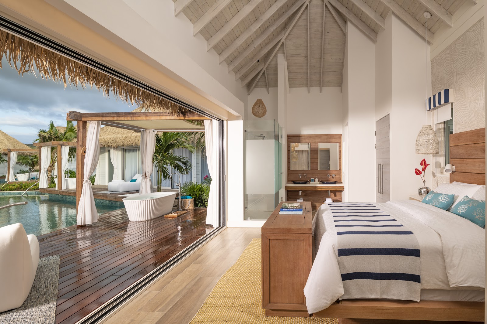 Bedroom with wooden bed and striped blanket opening through large glass doors to a private pool deck with soaking tub and ocean view.