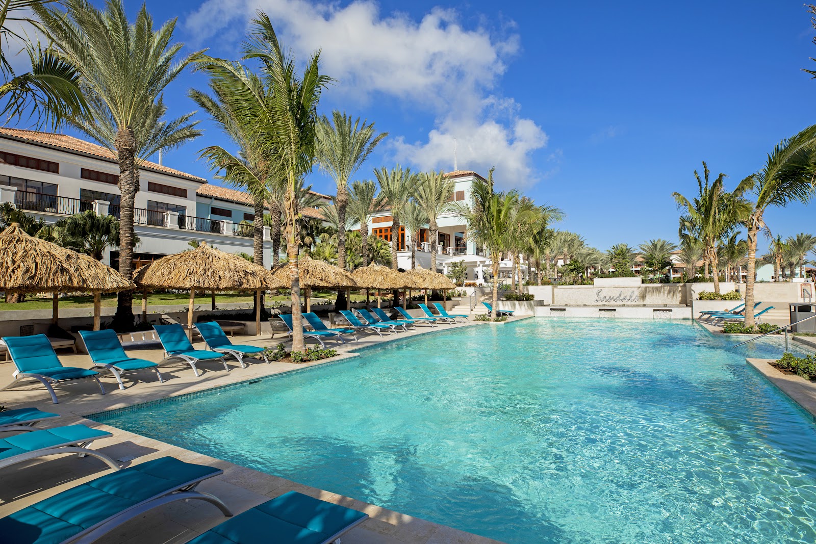 Turquoise pool floats rest on a tiled deck beside a clear swimming pool, with palm trees and manicured grounds visible beyond.