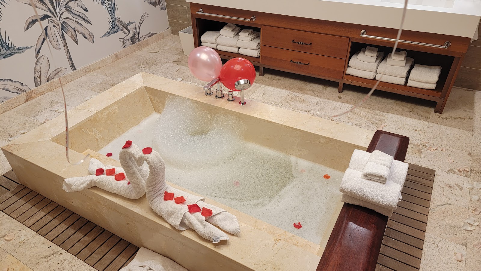 Sunken marble bathtub decorated with white towel swans and red rose petals, flanked by pink and red balloons and folded towels.