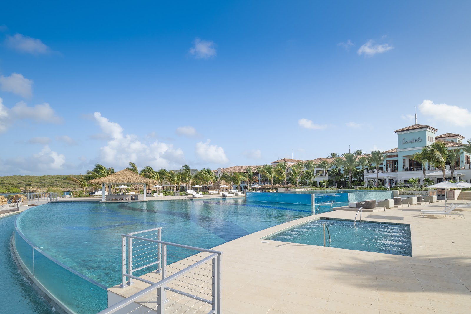 Curved lagoon-style pools with sun loungers and a turquoise main pool at Sandals Royal Curaçao, with a multi-story resort building and palm trees beyond.