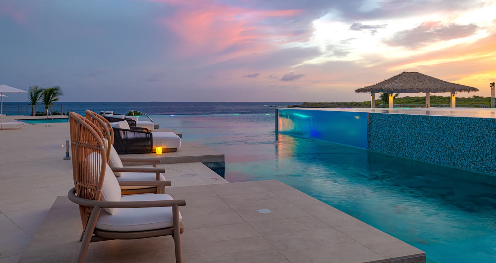 Lounge chairs line the infinity pool at Sandals Royal Curaçao at sunset, with the ocean and sky painted in pink and orange hues beyond.