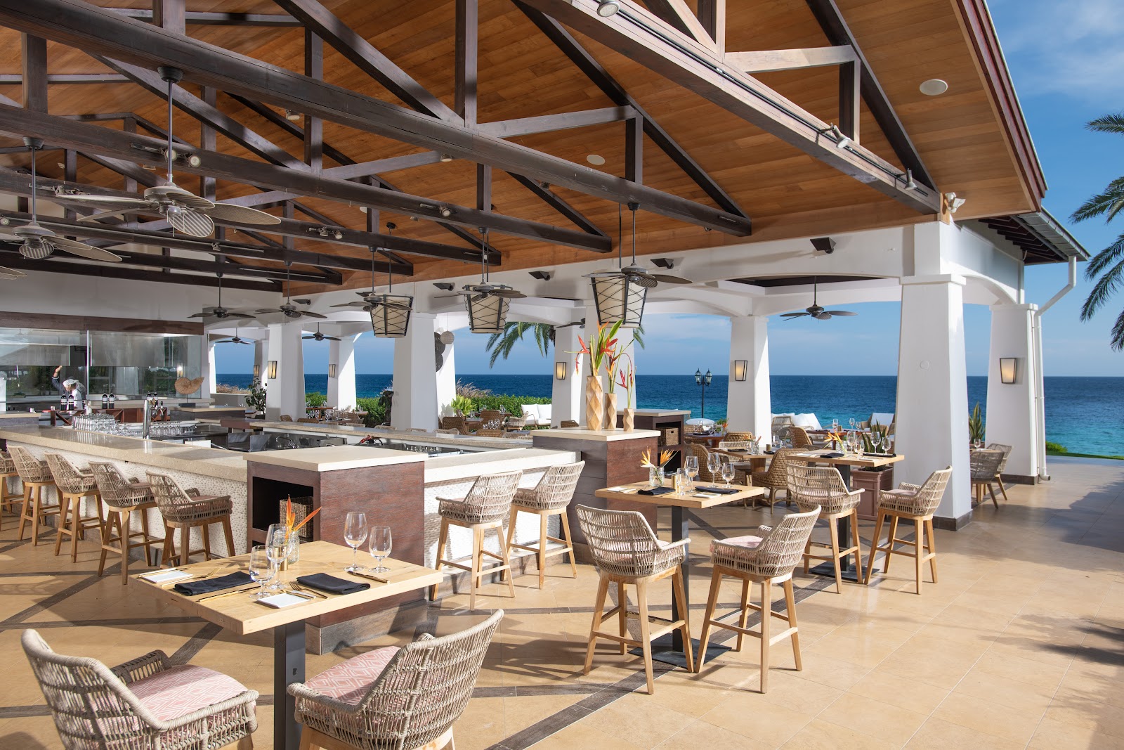 Beach lounge area with pallet furniture, palm trees, and turquoise water at Sandals Royal Curaçao, with a boat and hillside in the distance.
